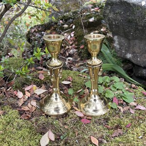 May include: Two brass candlesticks with a detailed design, set outdoors on moss and fallen leaves. The candlesticks have a flared base, a central decorative element, and a cup-shaped top for holding candles. The background features greenery and a large rock.