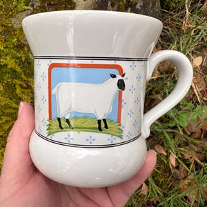 May include: White ceramic mug with a sheep illustration. The mug features a rounded base and a curved handle. The sheep is white with black legs and a black face, set against a blue and orange background with floral accents.