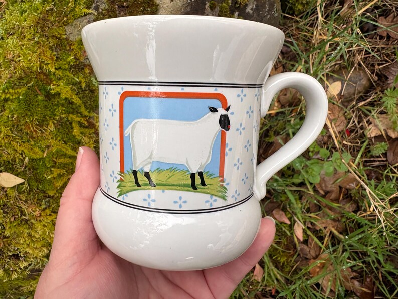 May include: White ceramic mug with a sheep illustration. The mug features a rounded base and a curved handle. The sheep is white with black legs and a black face, set against a blue and orange background with floral accents.