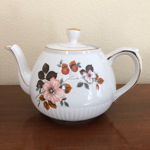以下が含まれることがあります： ピンクのバラ、茶色の葉、赤い実が描かれた花柄の白いティーポット。ティーポットの蓋と注ぎ口には金色の縁取りが施されています。