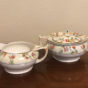 May include: A vintage cream-colored sugar and creamer set with a floral design in shades of orange, yellow, green, and pink. The set features a round sugar bowl with a lid and a creamer pitcher with a curved spout.