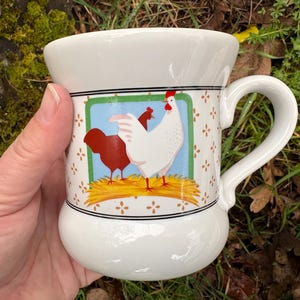 May include: A set of three ceramic mugs with a speckled, cream-colored glaze. Each mug features a different hand-painted design: a blue and green leaf, a yellow flower, and a red berry. The mugs have a simple, rounded shape.