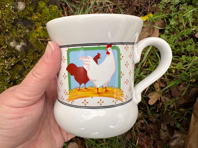 May include: A set of three ceramic mugs with a speckled, cream-colored glaze. Each mug features a different hand-painted design: a blue and green leaf, a yellow flower, and a red berry. The mugs have a simple, rounded shape.