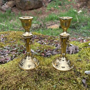 May include: Two brass candle holders with a classic design. Each holder features a flared top, a decorative rope-like detail, and a tassel accent. The candle holders are displayed on a bed of green moss.
