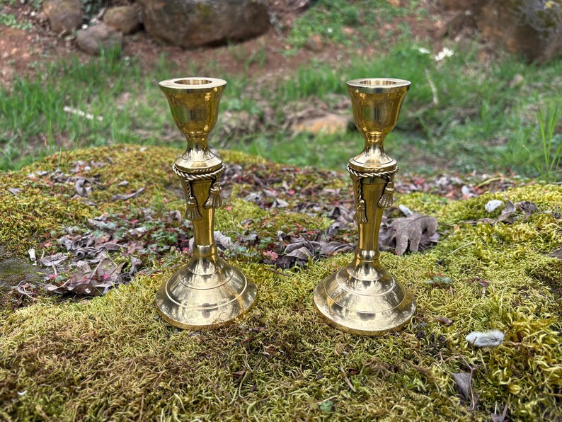 May include: Two brass candle holders with a classic design. Each holder features a flared top, a decorative rope-like detail, and a tassel accent. The candle holders are displayed on a bed of green moss.