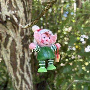 May include: A wooden Christmas elf ornament with a green tunic, pink ears, and a white and red hat. The elf has a painted face and holds a small bucket. The ornament is hanging from a tree branch against a blurred green background.