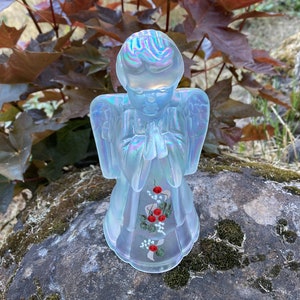 May include: A white iridescent glass angel figurine with painted red flowers and green leaves. The angel is praying with its hands clasped together.