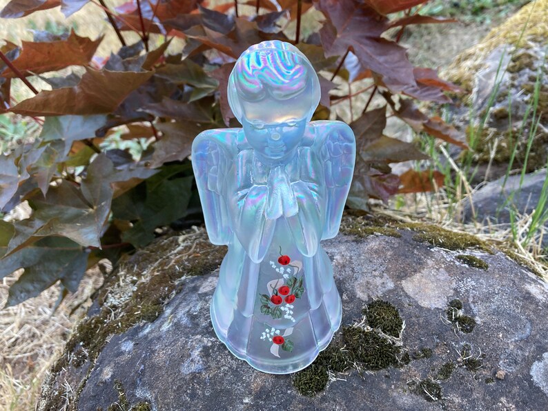 May include: A white iridescent glass angel figurine with painted red flowers and green leaves. The angel is praying with its hands clasped together.