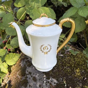 May include: A white porcelain teapot featuring a gold-colored handle, a gold-trimmed lid with a small finial, and a gold-colored spout tip. The body of the teapot is decorated with a gold-colored laurel wreath emblem surrounding the letter G.