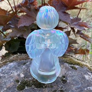 May include: A white iridescent glass angel figurine with wings outstretched. The angel is standing on a rock with moss.