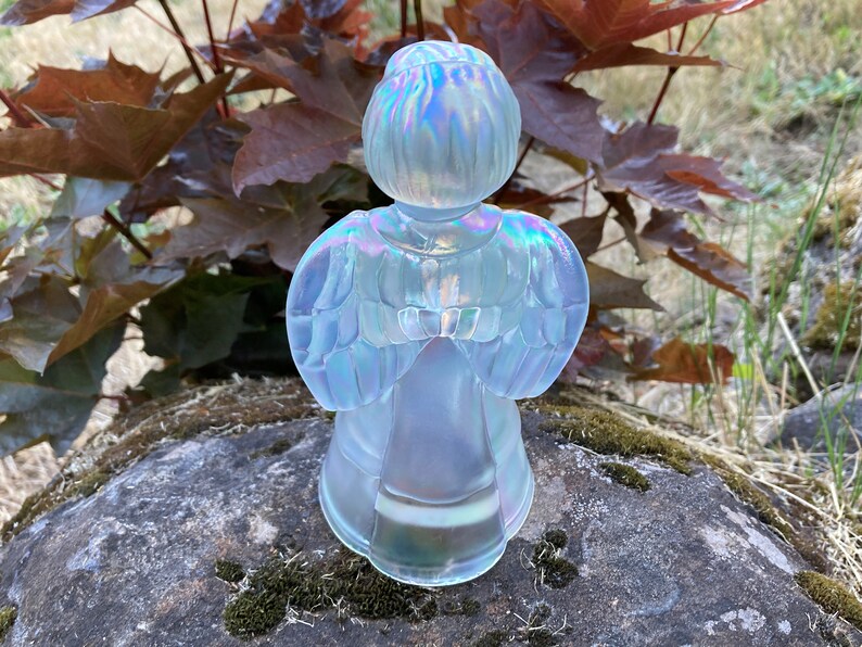 May include: A white iridescent glass angel figurine with wings outstretched. The angel is standing on a rock with moss.