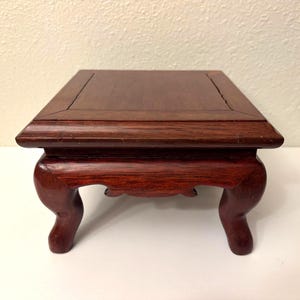 Può includere: Piccolo espositore quadrato in legno con una ricca finitura marrone-rossastra. Il supporto presenta un piano piatto con un bordo leggermente rialzato e gambe elegantemente curve. Ideale per esporre piccoli oggetti da collezione.