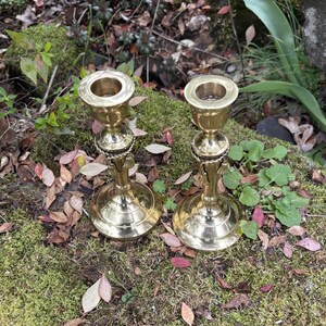 May include: Two brass candlesticks with a classic design. Each candlestick has a round base, a decorative stem, and a cup-shaped top to hold a candle. The candlesticks are displayed on a bed of moss and fallen leaves.