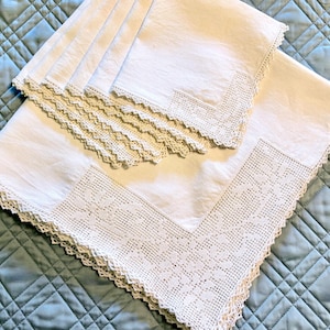 May include: A set of white linen napkins and a tablecloth with intricate crochet lace detailing. The napkins are neatly stacked, and the tablecloth is partially unfolded, showcasing the delicate floral patterns and scalloped edges.