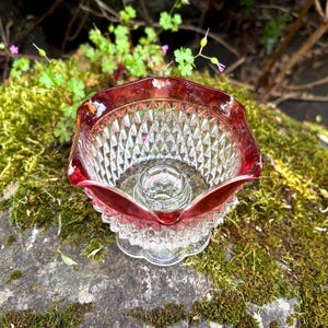 Può includere: Una ciotola vintage in vetro, con bordo rosso rubino e motivo trasparente a taglio diamante. La ciotola ha una base decorativa. La ciotola è probabilmente utilizzata per scopi decorativi.
