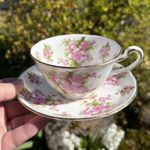 K&ouml;nnte beinhalten: Eine wei&szlig;e Teetasse und Untertasse mit einem floralen Muster aus rosa Bl&uuml;ten und gr&uuml;nen Bl&auml;ttern. Der Rand und der Henkel der Tasse sowie der Rand der Untertasse sind goldfarben verziert. Die Tasse und Untertasse werden von einer Hand gehalten.
