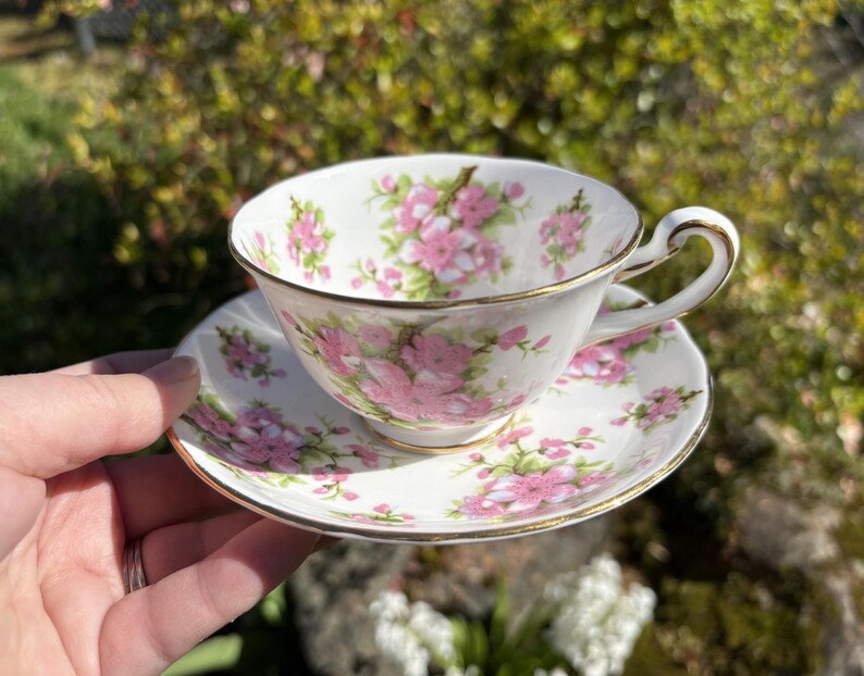 K&ouml;nnte beinhalten: Eine wei&szlig;e Teetasse und Untertasse mit einem floralen Muster aus rosa Bl&uuml;ten und gr&uuml;nen Bl&auml;ttern. Der Rand und der Henkel der Tasse sowie der Rand der Untertasse sind goldfarben verziert. Die Tasse und Untertasse werden von einer Hand gehalten.