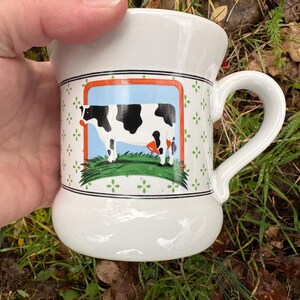 May include: White ceramic mug with a curved handle. The mug features a rectangular image of a black and white cow on a blue background, framed by an orange border. The mug has a decorative green floral pattern.