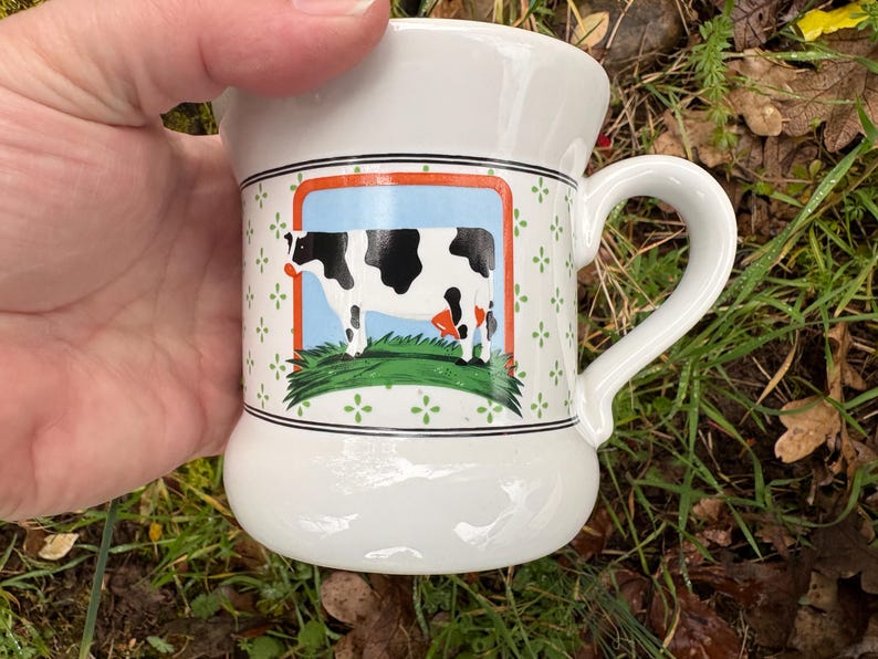 May include: White ceramic mug with a curved handle. The mug features a rectangular image of a black and white cow on a blue background, framed by an orange border. The mug has a decorative green floral pattern.