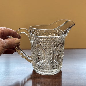May include: A clear glass pitcher with a diamond pattern and a handle. The pitcher is on a wooden surface.