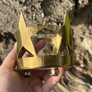 Puede incluir: Un portavelas de metal dorado con recortes de estrellas. El portavelas tiene forma cil&iacute;ndrica con tres peque&ntilde;os pies de bola. Los recortes de estrellas est&aacute;n espaciados uniformemente alrededor de la circunferencia.