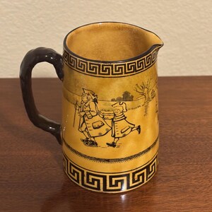 May include: A vintage ceramic pitcher with a brown handle and spout. The pitcher features a yellow background with black Greek key border designs and a black illustration of two figures walking. The interior is yellow.