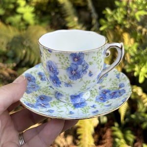 May include: A delicate teacup and saucer set. The white porcelain is adorned with a blue floral pattern and gold trim. The teacup has a curved handle and sits on a matching saucer. The set is designed for serving tea.
