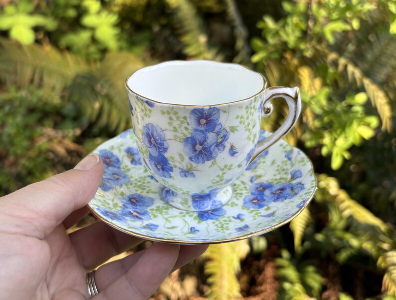 May include: A delicate teacup and saucer set. The white porcelain is adorned with a blue floral pattern and gold trim. The teacup has a curved handle and sits on a matching saucer. The set is designed for serving tea.