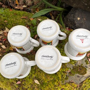 May include: Five white ceramic mugs with handles, arranged on a mossy surface. The mugs have a design around the middle and the text "Fender Country" on the bottom. The mugs are in a natural outdoor setting.