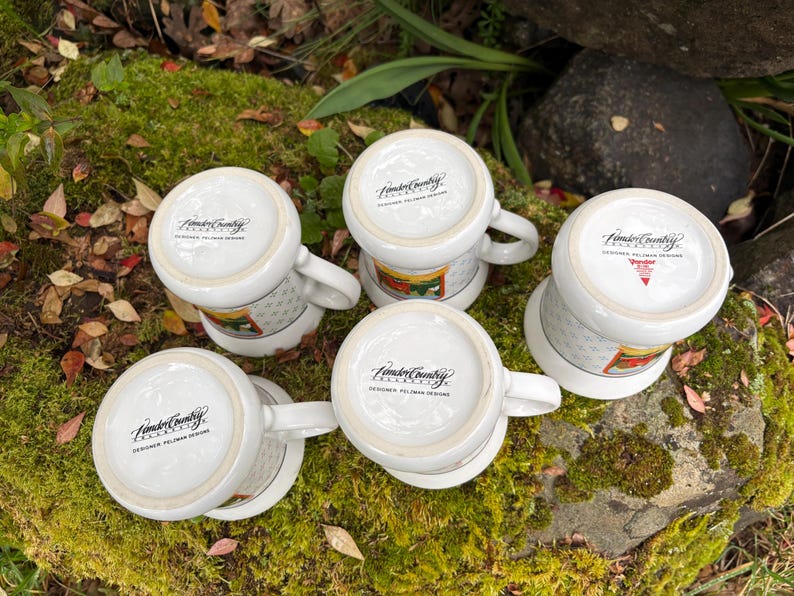 May include: Five white ceramic mugs with handles, arranged on a mossy surface. The mugs have a design around the middle and the text "Fender Country" on the bottom. The mugs are in a natural outdoor setting.