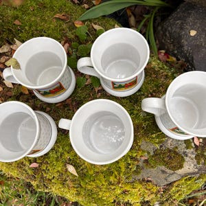 May include: A collection of six white ceramic mugs with a handle. Each mug has a decorative band with a colorful image. The mugs are arranged on a mossy surface with leaves and greenery in the background.