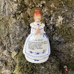 Op de afbeelding: Vintage keramieken beeldje van een meisje in een witte jurk met blauwe bloemmotieven, handen gevouwen in gebed. De jurk heeft de tekst "Dinner Prayer" en een gebed. Het beeldje heeft rood haar en staat tegen een mosachtige achtergrond.