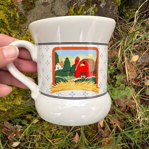 May include: White ceramic mug with a farm scene illustration. The mug features a handle and a decorative band with blue floral accents. The farm scene includes a red barn, green trees, and a field of hay.