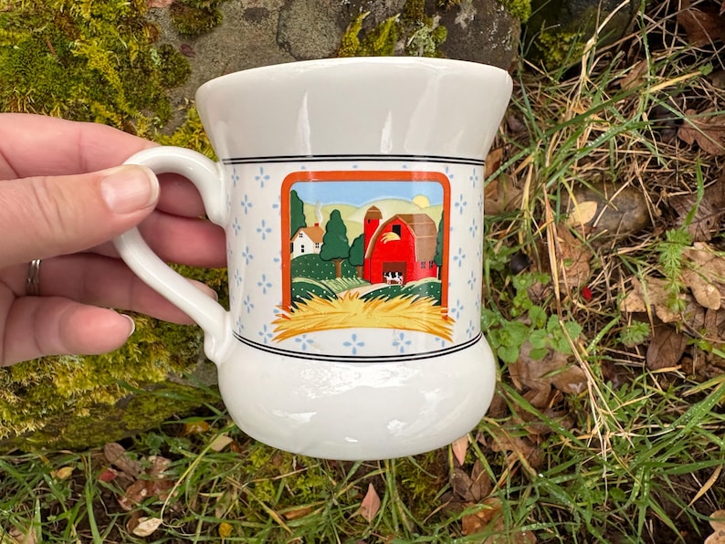 May include: White ceramic mug with a farm scene illustration. The mug features a handle and a decorative band with blue floral accents. The farm scene includes a red barn, green trees, and a field of hay.