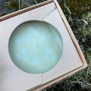 May include: A close-up of a light pink cardboard box with a circular cutout. The cutout is for a round object, such as a candle or a soap.