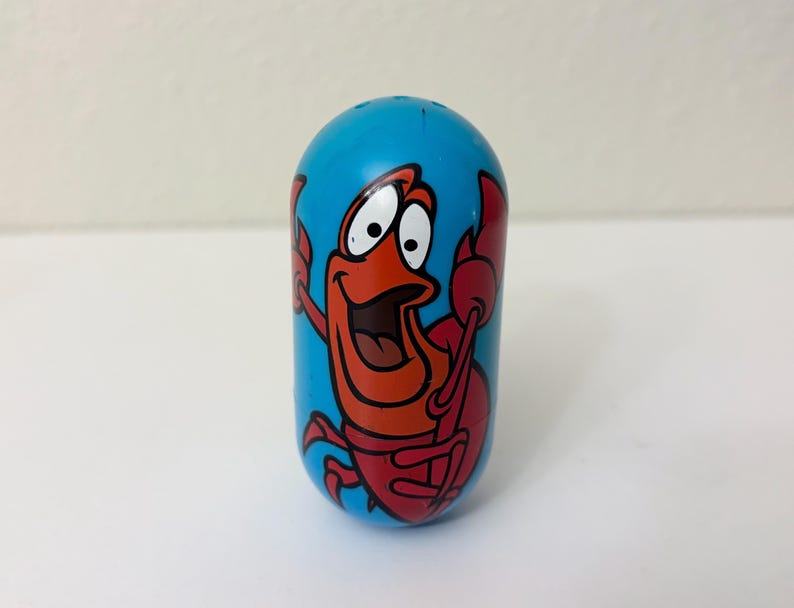 May include: A blue, oval-shaped toy featuring a cartoon crab character with a red body and claws, and a smiling face. The crab is set against a bright blue background.