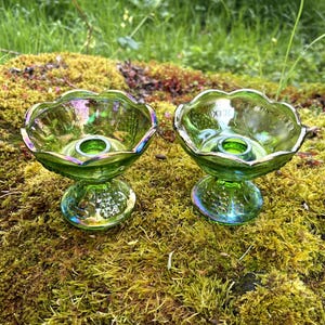 Puede incluir: Dos candelabros de vidrio verde con bordes y bases iridiscentes. Los candelabros tienen un borde festoneado y se asientan sobre una superficie musgosa. El vidrio tiene una ligera iridiscencia, reflejando múltiples colores.