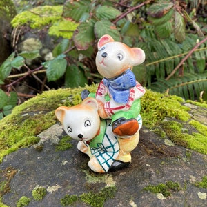 May include: Two ceramic bears, one riding on the other's back. The bear on top is wearing a red and white plaid shirt, green pants, and a blue scarf. The bear on the bottom is wearing a green and white plaid shirt. Both bears have painted eyes and noses.