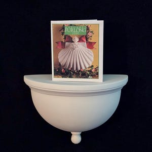 Puede incluir: Una estantería de pared blanca, semicircular, con una base decorativa redondeada y un pequeño pie redondo. Una tarjeta de felicitación con el texto "MARGARET FURLONG 2000" y un diseño de concha se muestra en la estantería.