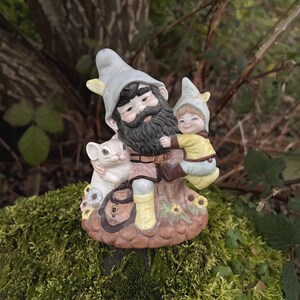 以下が含まれることがあります： ひげを生やしたノームが子供と白いマウスを抱いている、セラミック製の庭のノームの置物。ノームは灰色の帽子をかぶり、子供は黄色のシャツを着ています。ベースは花で飾られ、苔むした表面に置かれています。