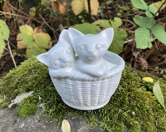 May include: A white ceramic ornament featuring two cats in a woven basket. The cats are detailed and close together. The basket has a textured, woven design. The ornament is set against green moss and foliage, creating a natural backdrop.