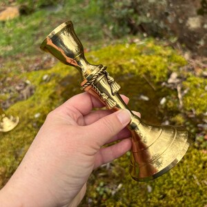 May include: A shiny, gold-colored candlestick with a flared base and a bell-shaped top. The candlestick has a decorative rope and tassel design around the center. Another smaller candlestick is visible in the background.