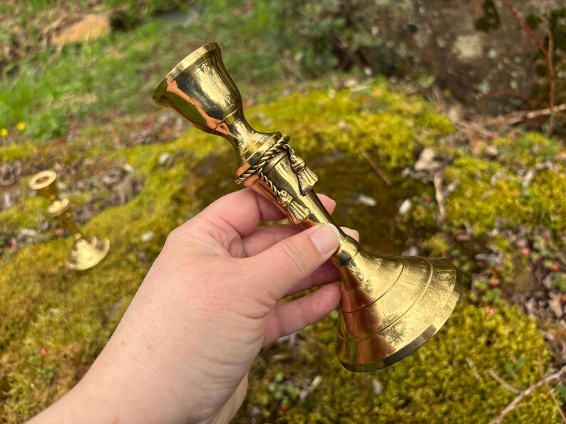 May include: A shiny, gold-colored candlestick with a flared base and a bell-shaped top. The candlestick has a decorative rope and tassel design around the center. Another smaller candlestick is visible in the background.