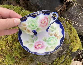 Puede incluir: Un juego de taza y platillo vintage con fondo azul cobalto y diseños florales pintados a mano. La taza tiene tres pequeños pies y detalles dorados.