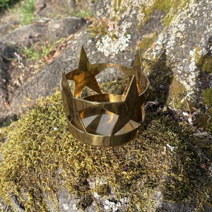 Puede incluir: Un portavelas de metal dorado con forma de corona y recortes de estrellas. El portavelas est&aacute; sobre un lecho de musgo verde y rodeado de rocas. El portavelas tiene una base circular y puntas en forma de estrella.