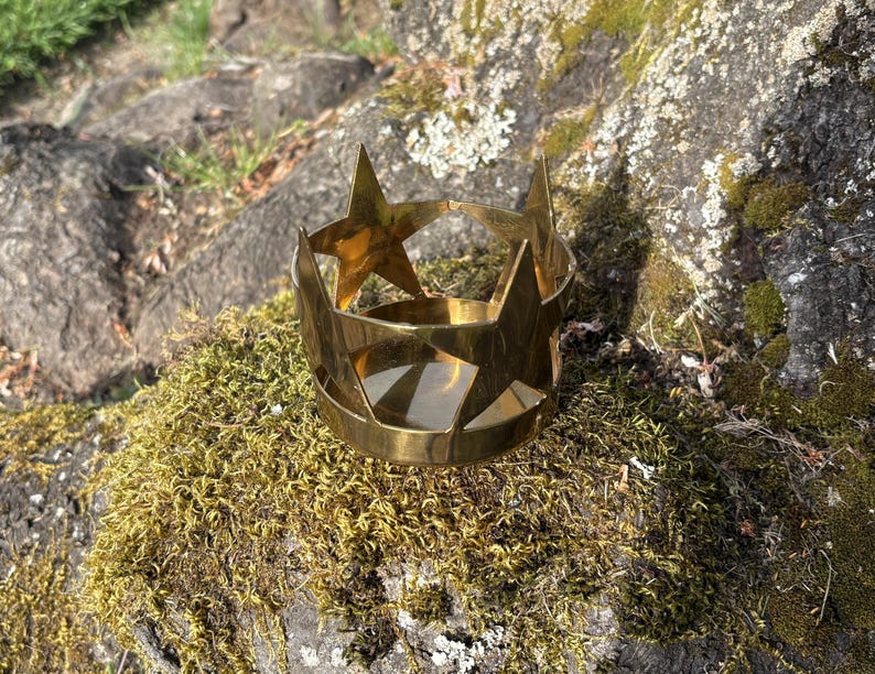 Puede incluir: Un portavelas de metal dorado con forma de corona y recortes de estrellas. El portavelas est&aacute; sobre un lecho de musgo verde y rodeado de rocas. El portavelas tiene una base circular y puntas en forma de estrella.