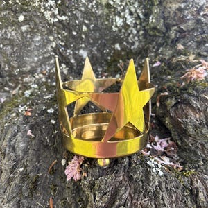 Puede incluir: Un portavelas de metal dorado con recortes de estrellas. El portavelas tiene una base circular y un dise&ntilde;o en forma de corona con detalles en forma de estrella. El objeto est&aacute; colocado sobre una superficie texturizada de color oscuro.