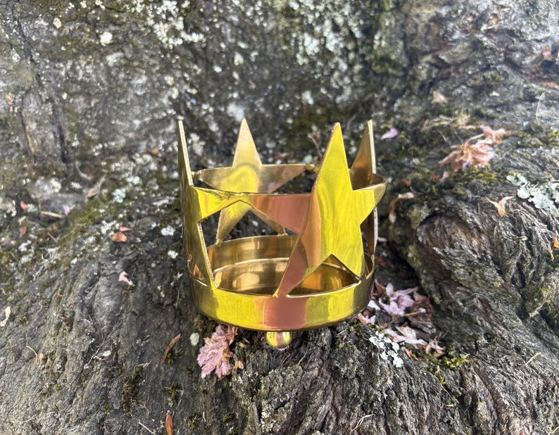 Puede incluir: Un portavelas de metal dorado con recortes de estrellas. El portavelas tiene una base circular y un dise&ntilde;o en forma de corona con detalles en forma de estrella. El objeto est&aacute; colocado sobre una superficie texturizada de color oscuro.