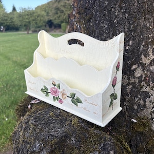 Puede incluir: Portacartas de madera color crema con diseño de rosas florales. El organizador tiene tres compartimentos y un asa en la parte superior. La parte delantera está decorada con rosas rosas y blancas y hojas verdes. Los lados también presentan ilustraciones de rosas.