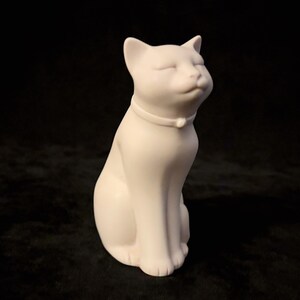 May include: A matte white ceramic cat figurine in a seated position. The cat has its head tilted upward with eyes closed in a serene expression and wears a simple collar with a small circular charm centered at the front of the neck.